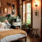 Cosy bedroom with soft lighting (lamp and wall sconce), many leafy plants, natural fibers/materials and textures, and earthy colours with a woman resting on the bed and looking out the window all lend to a hygge home