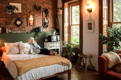 Cosy bedroom with soft lighting (lamp and wall sconce), many leafy plants, natural fibers/materials and textures, and earthy colours with a woman resting on the bed and looking out the window all lend to a hygge home
