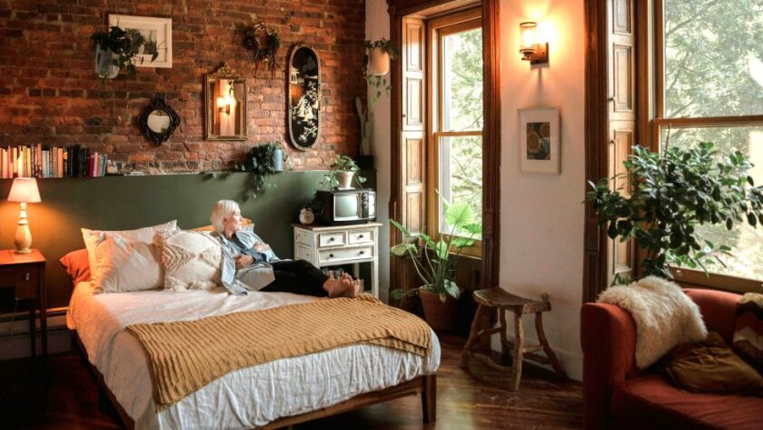 Cosy bedroom with soft lighting (lamp and wall sconce), many leafy plants, natural fibers/materials and textures, and earthy colours with a woman resting on the bed and looking out the window all lend to a hygge home