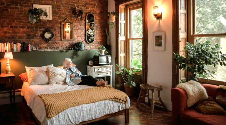 Cosy bedroom with soft lighting (lamp and wall sconce), many leafy plants, natural fibers/materials and textures, and earthy colours with a woman resting on the bed and looking out the window all lend to a hygge home