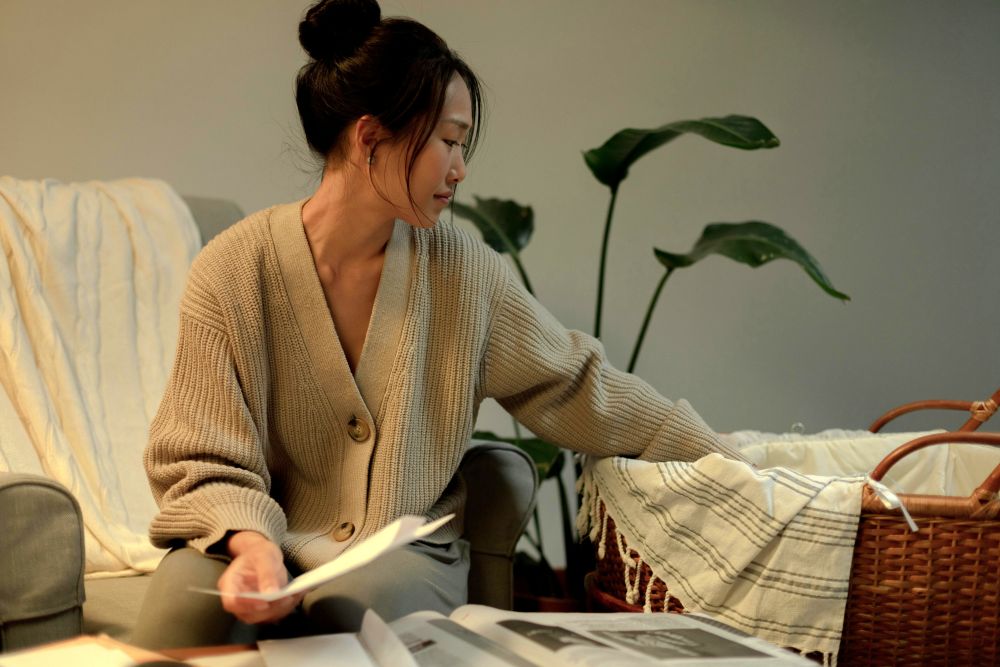 A woman in a cosy knit sweater sits next to her (unseen) sleeping baby in a wicker bassinette with blankets while reading the newspaper