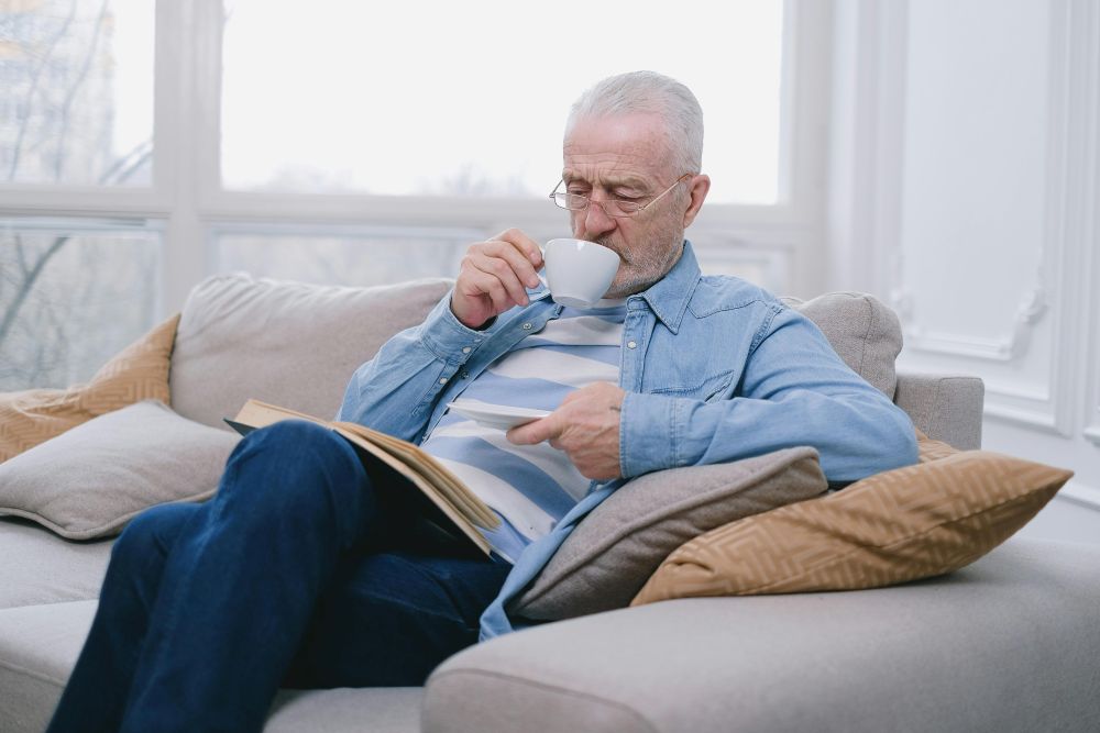 A man relaxes on a sofa with cushions while reading a book and sipping a mug of coffee