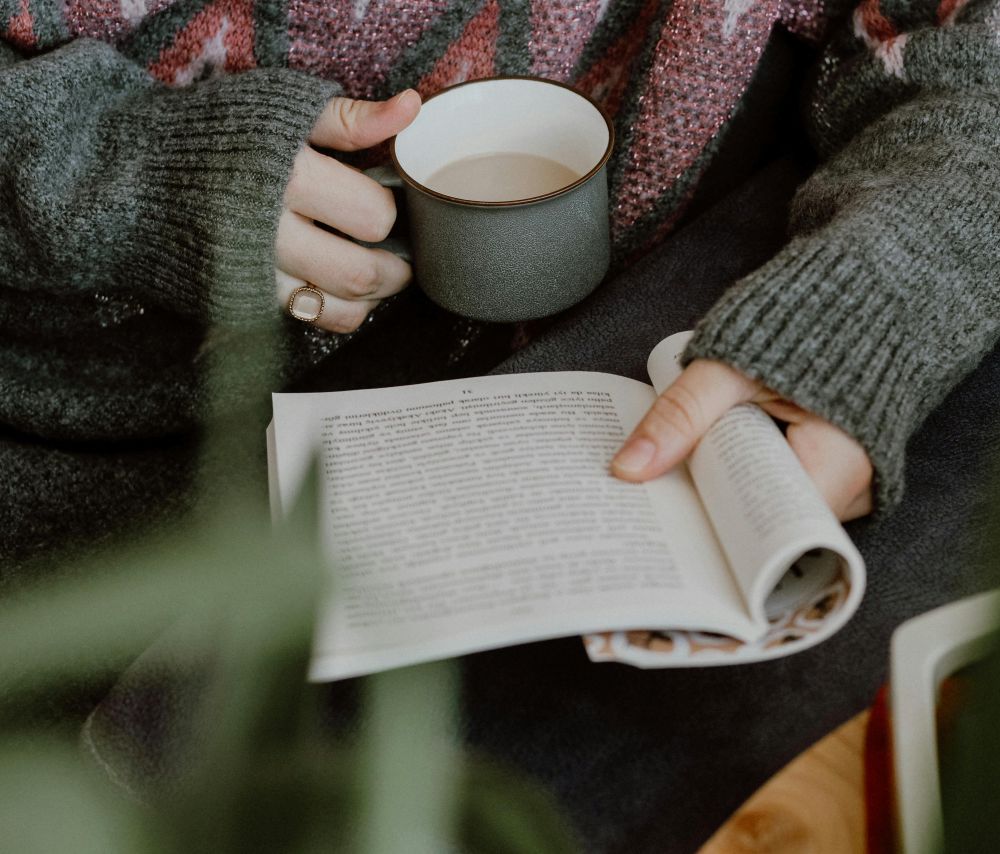 A woman wearing cosy sweaters holds a mug of coffee and reads an open book in her lap