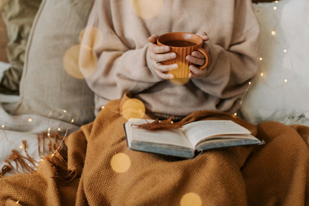 A woman in a baggy sweater holds a mug of coffee and sits under a blanket among cushions with string lights and an open book on her lap