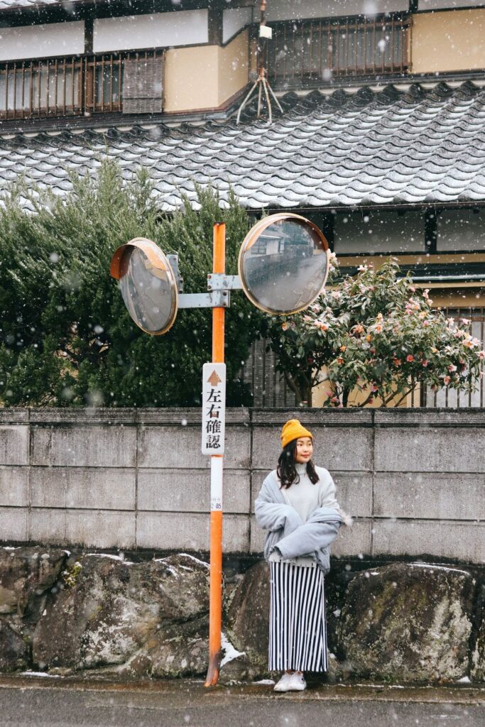 A woman wearing a long vertical black-and-white striped skirt, white sneakers, a large warm gray coat and mustard yellow winter hat stands by a street sign next to a wall lined with small trees/fence and house in Japan whilst snow falls