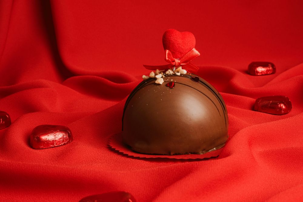 Close-up view of a perfectly round dome-shaped chocolate truffle with tiny nut crumbles on top as well as two metallic glitter hearts and a larger felt heart with a bow stuck on top (all in red), presented on red fabric. Chocolate is important not only during Valentine's Day but every time of year.