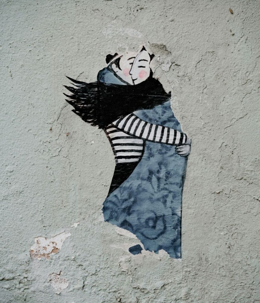 Photo of an artistic/graffiti-style painting on a cement wall in Lisbon of a man in a black and white striped shirt and a woman in a long blue gray dress with long black hair lovingly embracing