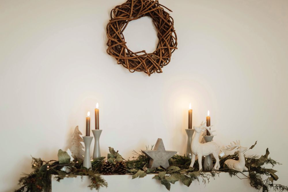 A simple and natural woven twig wreath hangs on a wall above a mantel covered in winter greenery, gray/white candlesticks with lit taper candles, and white/gray deer, star and evergreen tree figurines (a beautiful way to segue from Christmas into January winter decor)