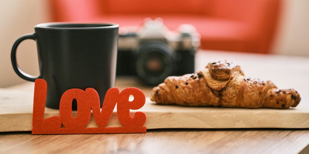 Close-up photo of February love: a felt red decoration reading 'Love' is propped against the side of a wood slab on top of which rests a black mug of coffee and a croissant in a cafe