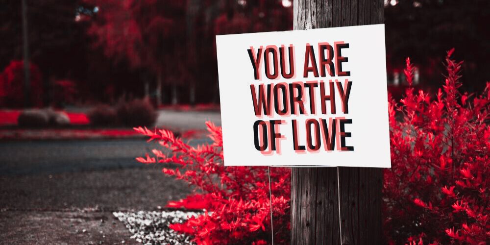 Close-up view of a sign stuck in the ground at the base of a wooden telephone pole/lamppost nearby some bushes and trees in background that reads 'YOU ARE WORTHY OF LOVE' and enhanced with a red filter, a beautiful reminder of February love for yourself and all others