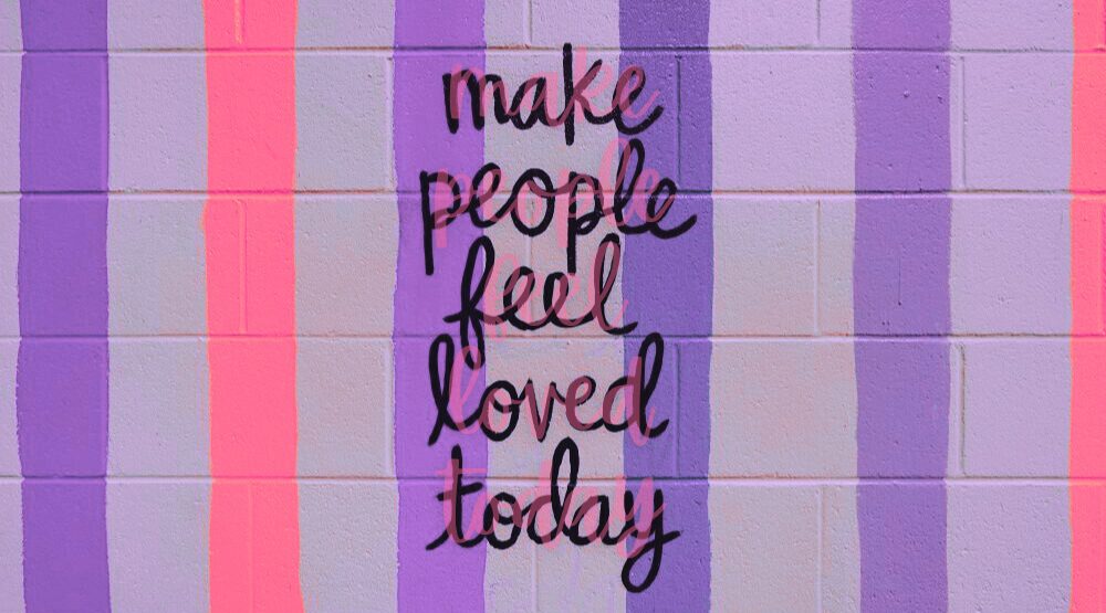 Photo of the phrase 'make people feel loved today' painted on striped cement block wall in Charlotte and colour-enhanced in periwinkle and bright peach/pink. A beautiful reminder of what February love is all about: loving others and ourselves