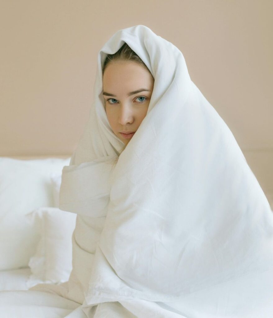 A girl wrapped up in a thick white comforter over her head sits in bed, reluctant to get out (something we all experience in cold January)