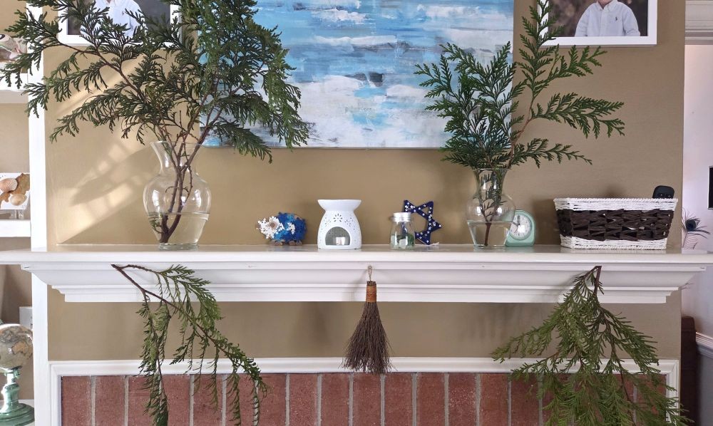 Large evergreen clippings fill two glass vases atop a fireplace mantel among other blue and white decor in front of a blue and white painting, and more evergreen branches hang from the mantel below