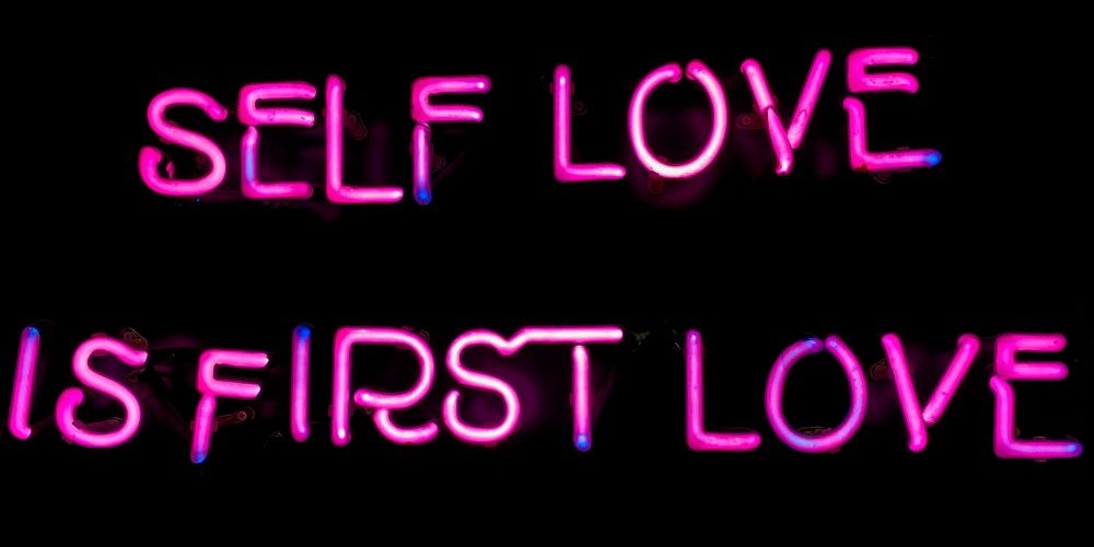 Close-up photo of a lit pink neon sign of letters reading 'SELF LOVE IS FIRST LOVE' against black background