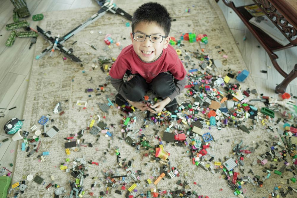 A young boy smiles while sitting in the middle of a rug with hundreds of tiny Lego pieces scattered about