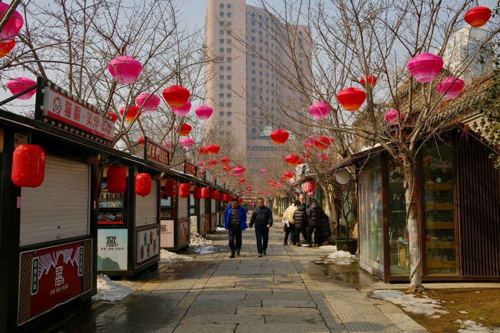 View of people walking down a stone footpath between various stalls with skyscrapers in background and trees lining walkway with hanging pink and red Chinese lanterns