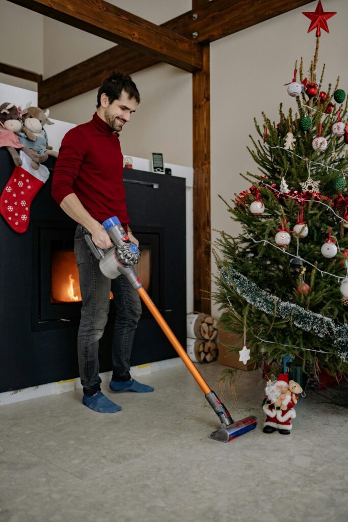 A man stands in front of a fireplace near a Christmas tree whilst vacuuming the floor