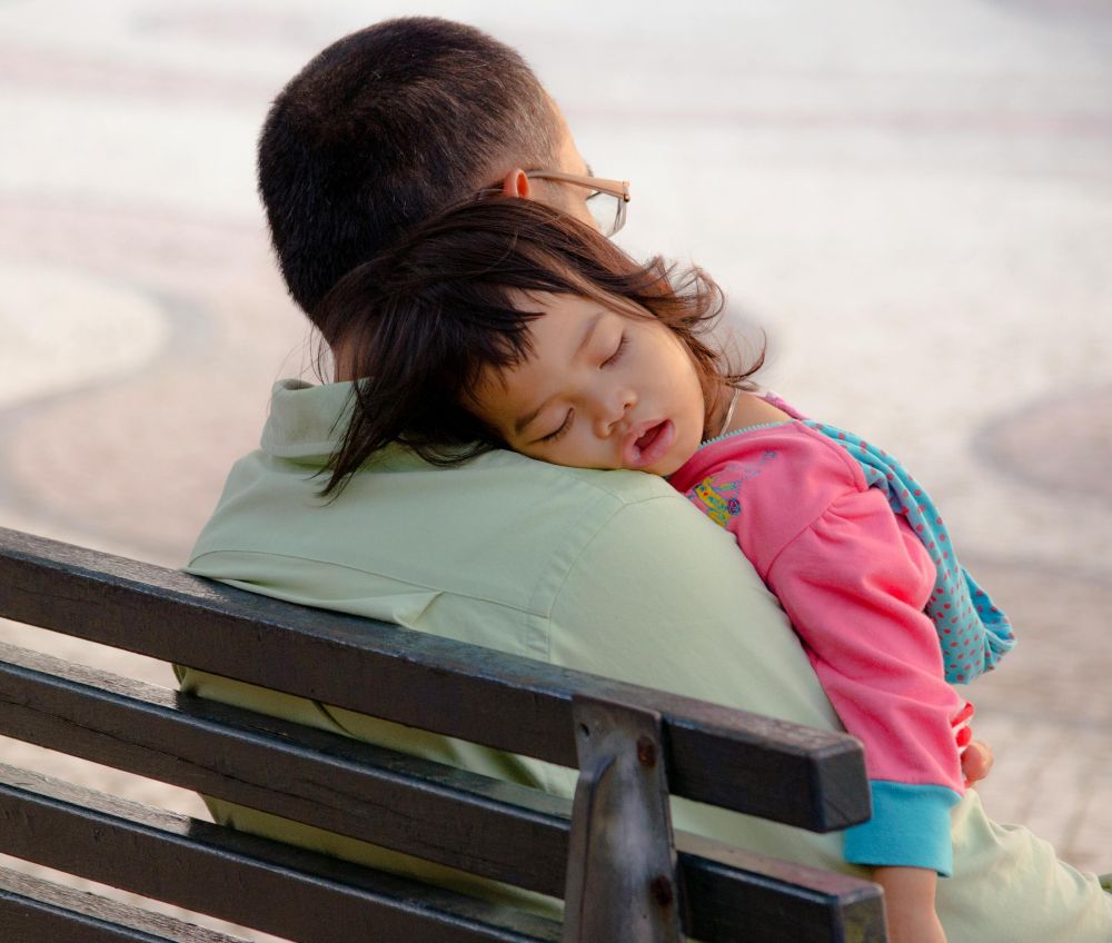 A little girl peacefully sleeps on the shoulder of her father sitting on a park bench