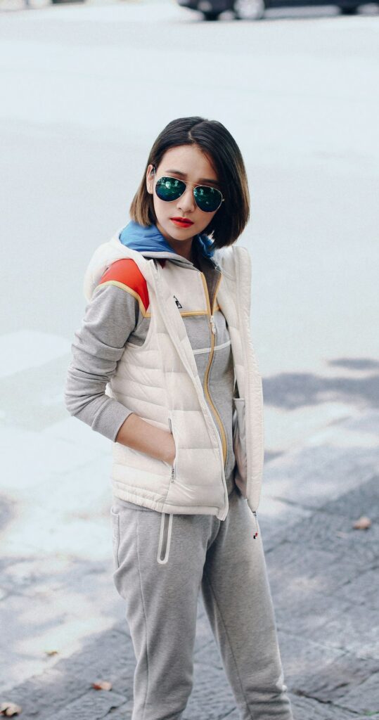 A young woman wearing a gray sweatsuit with blue and red block colour pops near the collar/hood with aviator sunglasses and a white puffer vest sports casual February fashion
