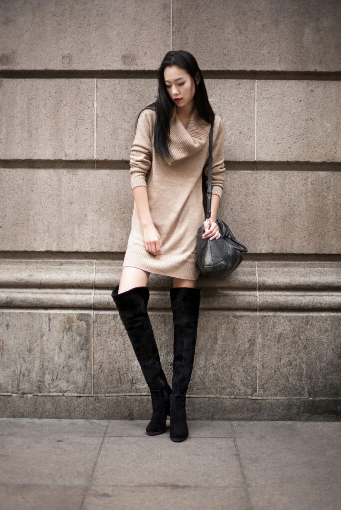 A woman in warm beige cowlneck dress and knee-high black boots leans against an exterior gray structure, a sleek February fashion look in neutral tones