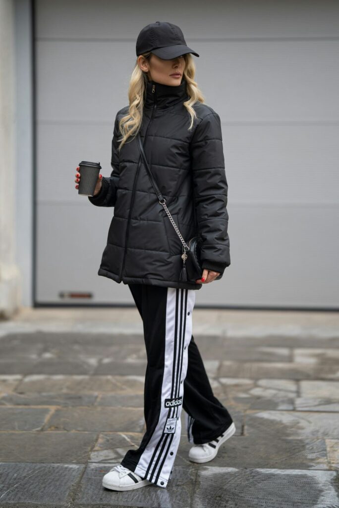 A woman wearing snap-up black and white Adidas track pants with black and white Adidas trainers wears a sleek black puffer coat, black purse and black ballcap while holding a black portable paper coffee cup