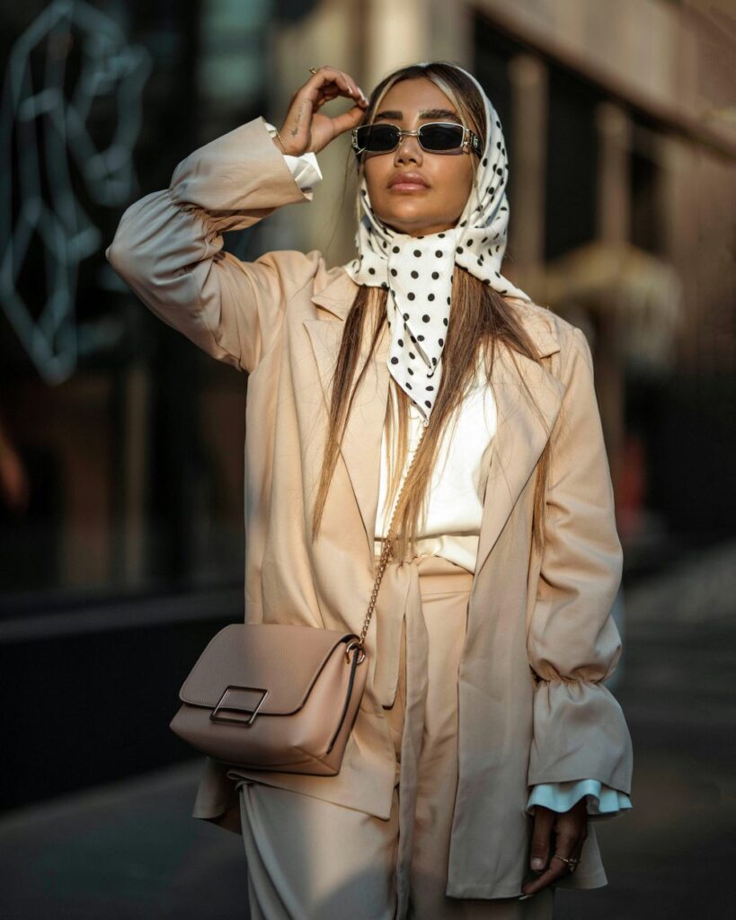 Fashionable woman in a tan jacket cinched at bell wrists and a polka dot headscarf tied around her head and neck with a beige purse and sunglasses