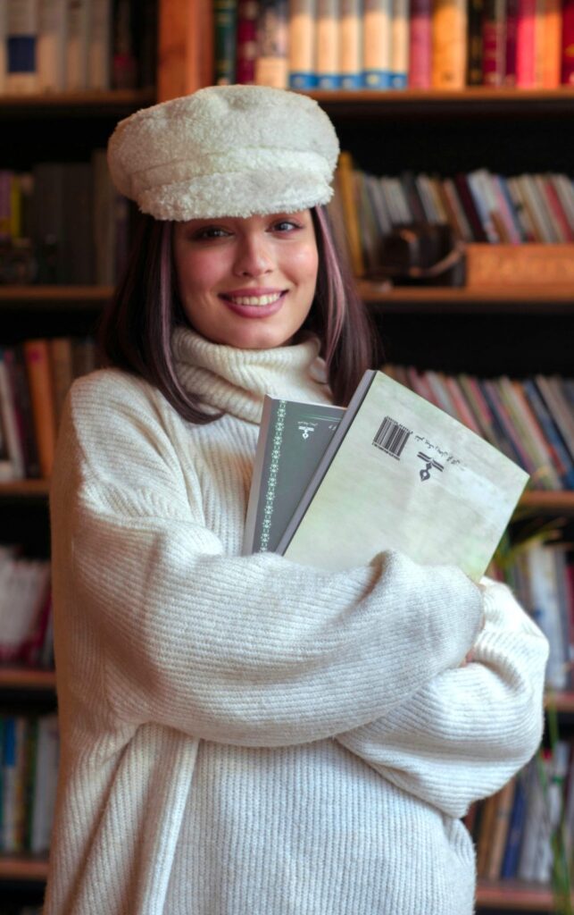 A smiling woman holes books and wears a soft oversized cream-coloured ribbed turtleneck sweater and matching cream wool hat