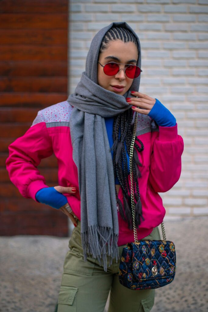 A woman poses in a bright pink jacket over electric blue sleeves covering wrists, red-tinted sunglasses, and gray scarf wrapped over her head and neck with a colourful purse over her shoulder