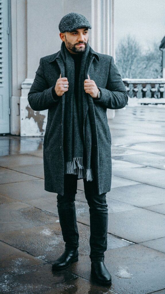 A man poses holding the lapels of a long gray overcoat with a matching fringed gray scarf, gray scally cap, black pants and black shoes