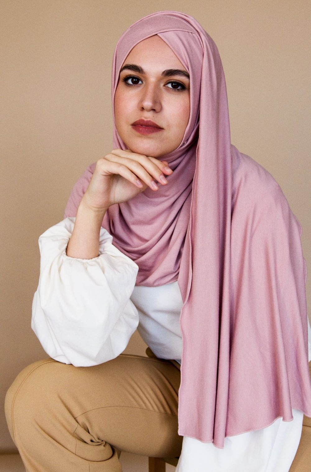 A woman poses in pale pink hijab that drapes down her shoulder, tan slacks, and a white blouse with poofy sleeves