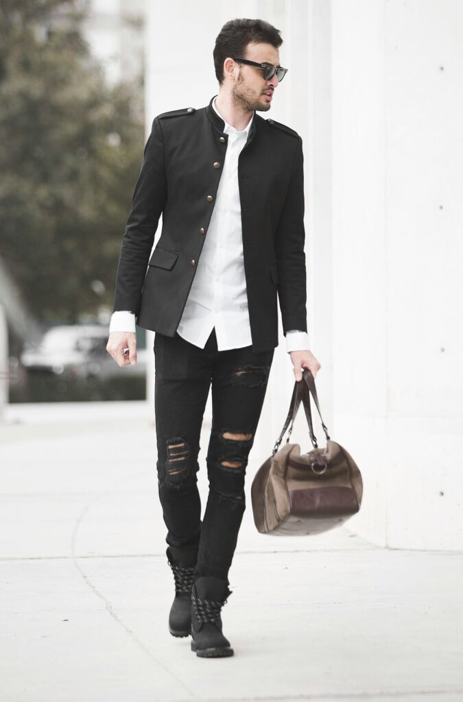 A man dressed business casual on top and punk rock on bottom embodies clever black and white February fashion and wears a white button-up shirt under a smart black business coat with gold buttons, skinny black jeans with intentional tears, and black boots and sunglasses whilst walking and carrying a large brown duffle bag