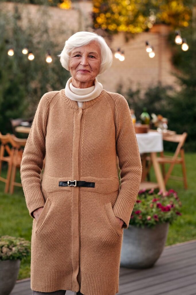 A smiling woman poses in a long tan zip-up sweater overtop of a soft cream turtleneck in a nicely landscaped garden and patio