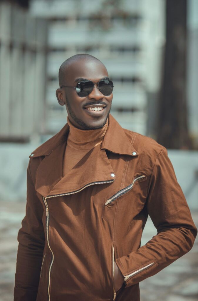 A smiling man in sunglasses poses in a muted tangerine turtleneck under a light brown jacket