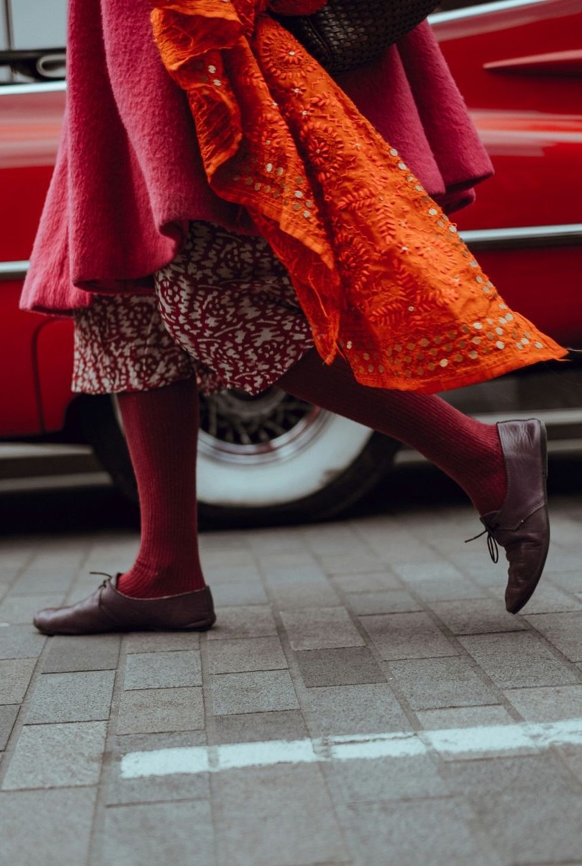 The lower half of a woman walking along a brick street in London showing dark red tights, maroon leather shoes, a long black and white skirt, long wool pink coat, and trailing bright orange scarf