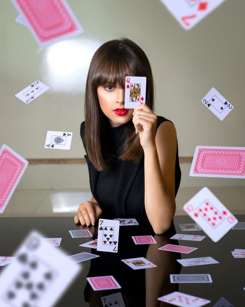 A woman sits at a table in a sleeveless black shirt holding a queen of diamonds playing card in front of one eye while other cards from the deck are strewn about the table and some flying midair