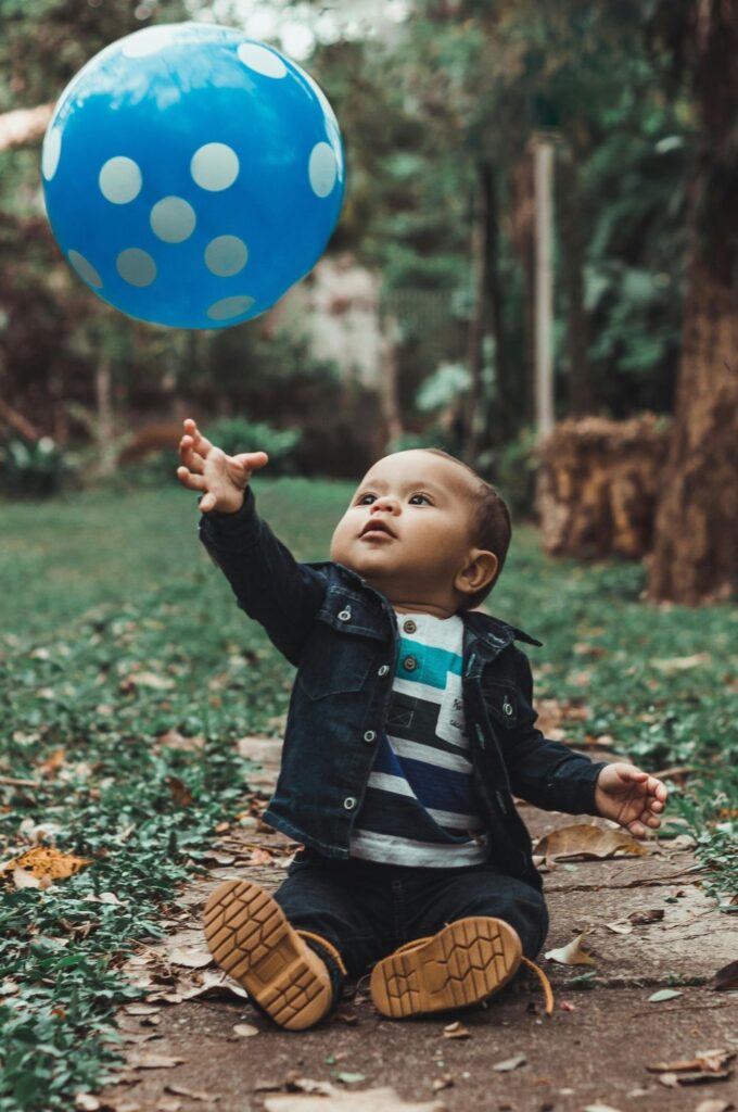 A baby boy sits on a footpath surrounded by grass and trees in the background with a big blue ball with white polka dots suspended in the air above his hand (photographed mid-throw)
