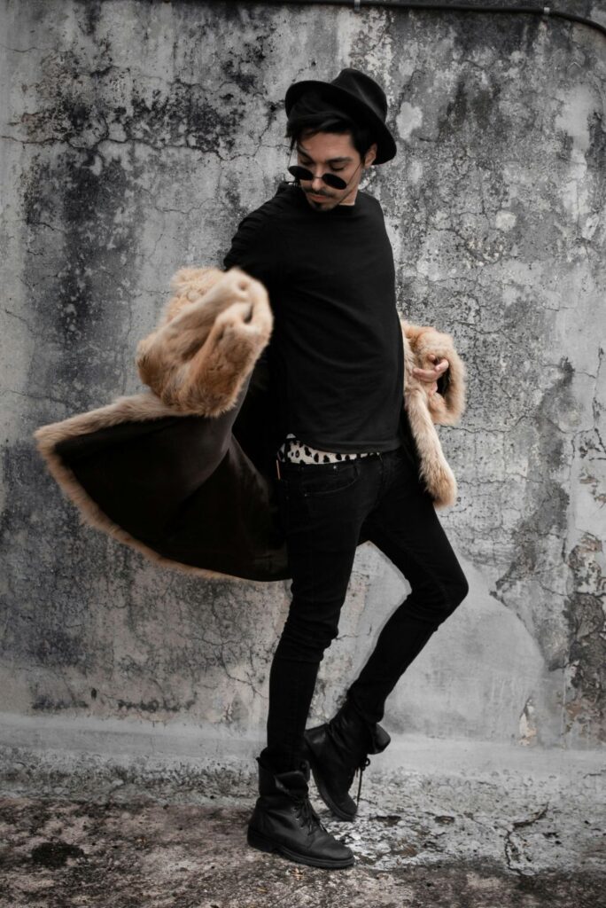 A young man poses mid-stride wearing black skinny jeans, black boots, a black sweater over a dalmatian-print shirt underneath, a black bowler hat and sunglasses, and a thick tan faux-fur cut for February fashion in action