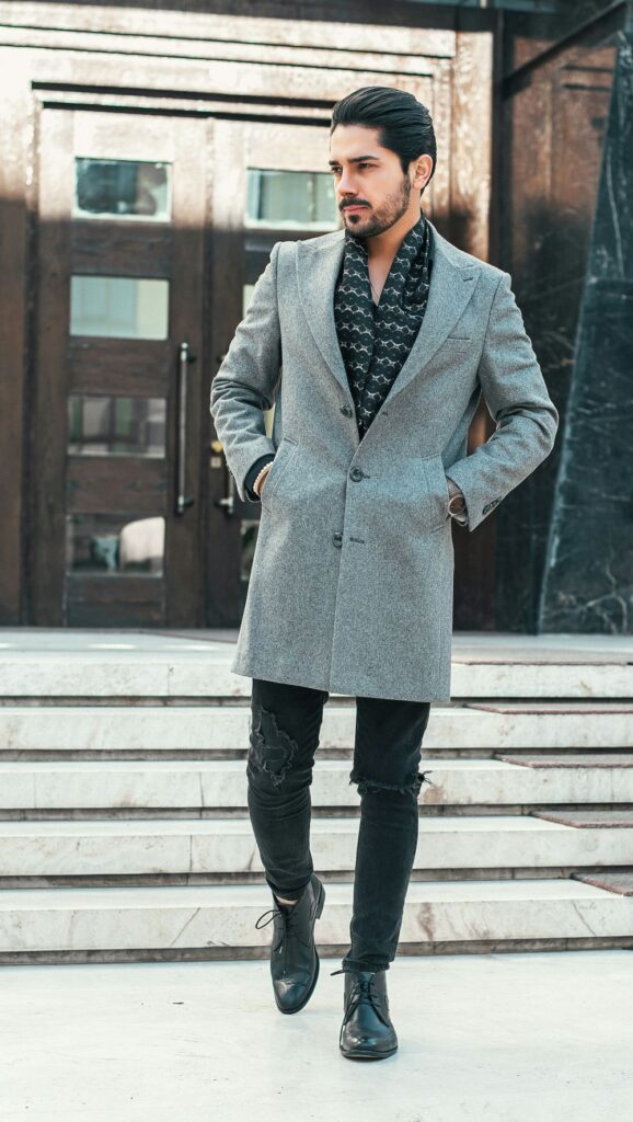One February fashion trend is pairing ripped jeans with classy taste: a man in Iran wears a smart gray overcoat over a patterned black scarf with ripped black jeans and black dress shoes in