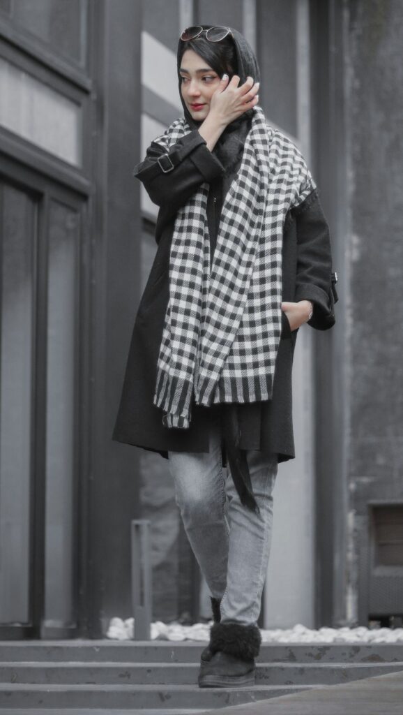 A woman walks beside a building wearing a long black overcoat, gray scarf over her head and a black and white chequered scarf draped over her shoulders with black sunglasses pushed up her head