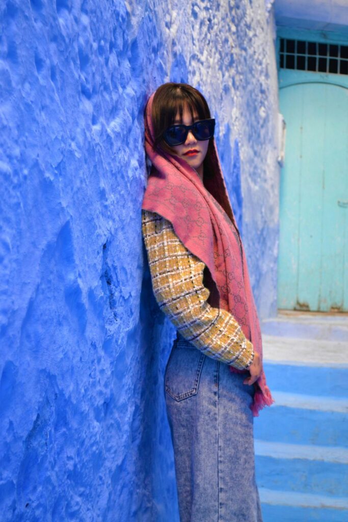 A woman leans against a blue textured stucco wall outside steps in Chefchaouen, Morocco wearing retro jeans, a mustard yellow and white tweed plaid jacket, sunglasses, and a patterned pink scarf draped over her head