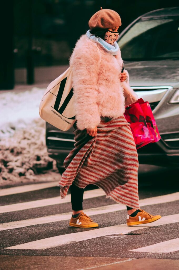 A woman crosses the street wearing a tan beret, fluffy feathery light pink coat, tan and black patterned face mask, long red and gray striped skirt, black leggings, and mustard yellow sneakers whilst carrying a large hot pink bag and a large white canvas bag with black print