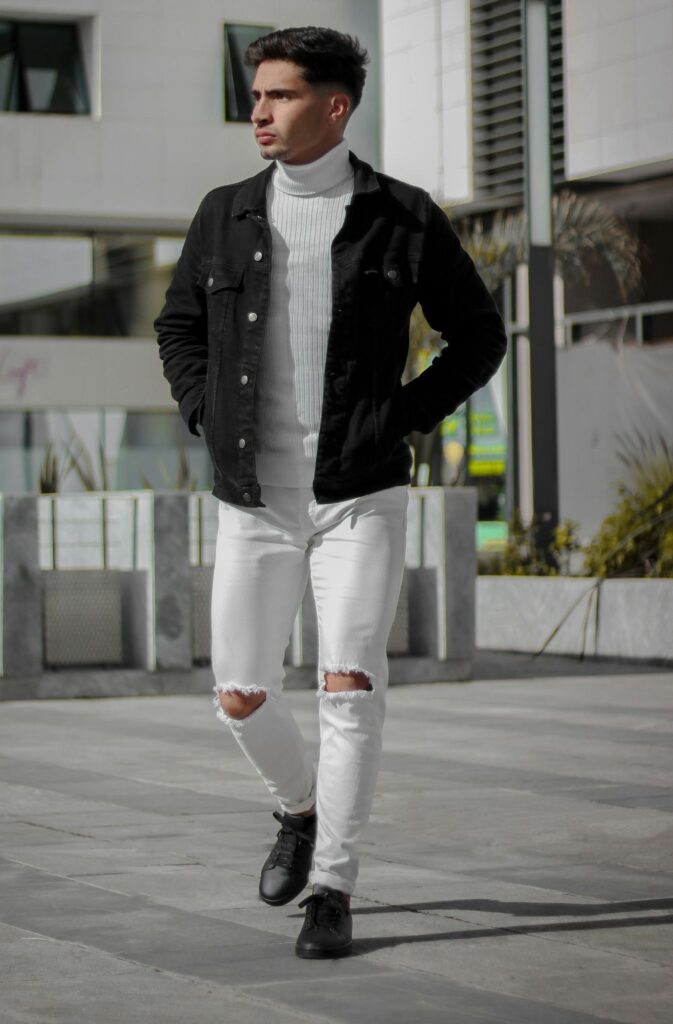 A man walks across a courtyard wearing a chic white ribbed turtleneck under a black denim jacket with black dress shoes and white jeans with holes at the knees: the trendy juxtaposition seen in February fashion pairing ripped jeans and dressy clothes