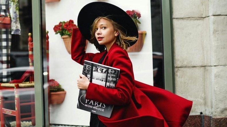 Woman in big black hat and big red coat walking down boutique-lined avenue holding a book about Paris