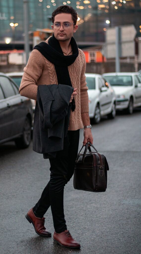 A man crosses a street in Tehran wearing a camel-coloured cable-knit sweater, black pants, brown dress shoes, and a black scarf draped over his neck while holding a gray overcoat over one arm and carrying a leather bag in the other. He embodies classic February fashion with neutral tones and smartly fitted winter staples