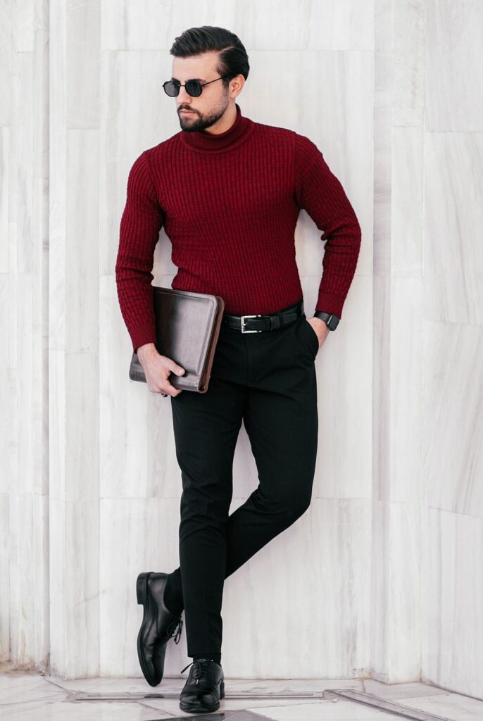 A chicly-dressed man stands against a marble wall in Iran wearing fitted black pants, black dress shoes, sunglasses, and a maroon turtleneck: a classy February fashion statement