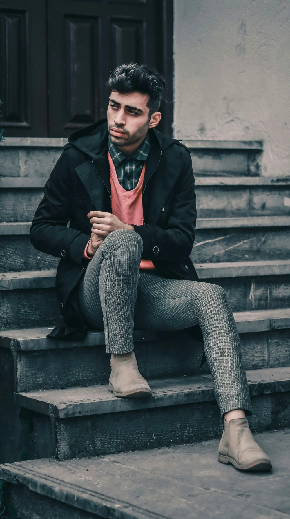 A man poses on steps outside a building in Rasht, Iran wearing a plaid button-up collared shirt under a coral-coloured shirt (colour pop) and black overcoat with pinstriped skinny pants and tan pull-on boots, for layered February fashion