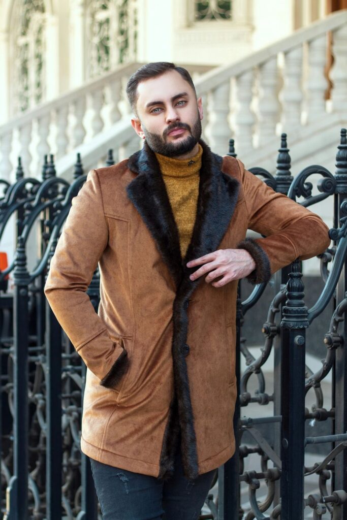 A man poses beside a wrought iron fence wearing a mustard turtleneck sweater under a tan suede coat lined with black fur lapels and cuffs