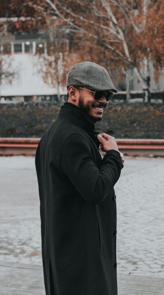 A man smiles in a long black overcoat with gray scally cap and sunglasses in New York