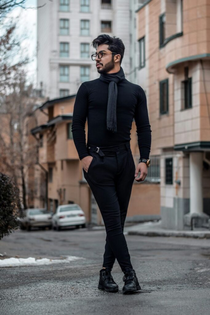 A man wearing fitted black pants, a fitted black turtleneck, black-framed glasses, black ankle boots and a dark gray scarf poses on the street