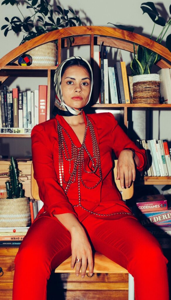 A woman lounges on a wooden chair in front of a busy bookshelf lined with stacks of books and houseplants wearing a stylish tomato red suit, black and white scarf tied over her head, black choker, and a large metal beaded statement necklace with waist-length layers - February fashion in fitting red